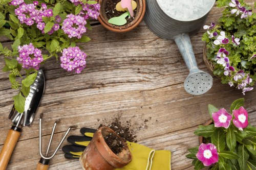 A wooden garden table with visible natural grain and weathered texture, displaying gardening tools and plant arrangements. There are potted flowering plants with purple and pink blossoms, including one with variegated green and white leaves. Gardening tools such as a small hand rake, a trowel, and a garden fork with wooden handles are placed on or near the table, alongside a small clay pot with soil, a yellow gardening glove, and a metal watering can with a long spout. The scene suggests an outdoor gardening space in mild weather, suitable for general gardening activities like planting, potting, and preparing flower beds, with natural lighting highlighting the vibrant colors of the flowers and the rustic surface of the wooden table. This setting aligns with gardening services offered by Landscapers St Johns Wood, reflecting attention to detail and garden maintenance in the local area near St Johns Wood, London.