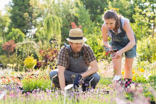 In a bright and lush garden, two women are engaging in gardening activities amidst a variety of colorful flower beds and greenery. The woman on the left, sitting on the grass, is wearing a straw hat, a checked shirt, and gardening gloves, focused on tending to the flowers or plants in front of her. The woman on the right, standing and leaning slightly forward, is dressed in a sleeveless top and denim shorts, holding garden tools and demonstrating careful pruning or planting. The garden features well-maintained grass, vibrant flowering plants with pink, yellow, and purple blooms, and a backdrop of mature trees and shrubs providing natural shade and privacy. The scene is set on a partly sunny day, with dappled sunlight illuminating the outdoor space, reflecting a healthy and inviting environment typical of a residential garden in St Johns Wood. This outdoor scene showcases a peaceful, cultivated garden area that could benefit from professional landscaping services offered by Landscapers St Johns Wood, emphasizing natural landscaping and garden maintenance tailored to local outdoor spaces.