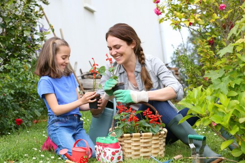 A woman and a young girl are gardening together in a lush, well-maintained outdoor garden space, with various green shrubs, flowering plants, and trees surrounding them. The woman, dressed in a grey jacket and green gardening gloves, is smiling while holding a small potted plant with red flowers, and she appears to be in the process of planting or tending to it. The girl, wearing a blue T-shirt and denim shorts, is assisting with the gardening task, holding the plant pot with both hands. On the grass nearby, there are gardening tools including a red watering can, a small garden spade, and a basket filled with gardening supplies, indicating active maintenance of the garden environment. The scene is set under natural daylight, suggesting a clear weather day, and the overall appearance of the garden features vibrant greenery, flowering plants, and a tidy, welcoming outdoor space typical of residential gardens in St Johns Wood, UK, maintained by professionals such as Landscapers St Johns Wood.