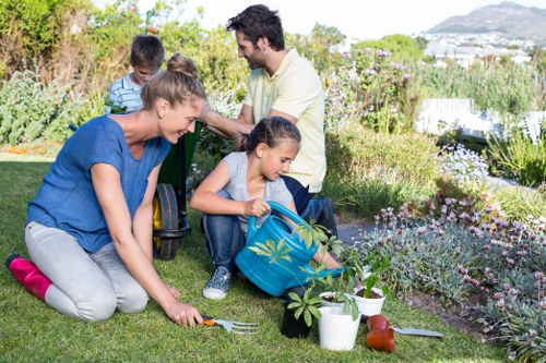 A family is tending to a well-maintained garden in a residential outdoor space, featuring lush green grass, various flowering plants, and garden beds bordered with natural soil and small paving stones. The foreground shows a woman planting a small shrub, while a young girl in a backpack is watering nearby plants with a plastic watering can. A man and a boy in a striped shirt are in the background, engaged in conversation amidst the garden's foliage, which includes flowering bushes and mature trees providing shade. The garden area appears tidy and cultivated, with gardening tools such as a trowel and watering cans visible, indicating active maintenance. Soft natural light suggests a bright, clear day, and distant views of the local landscape hint at a suburban setting, possibly within the St Johns Wood area in London. The scene exemplifies sustainable gardening practices, underlining environmental responsibility common to professional landscaping services offered by Landscapers St Johns Wood, reflecting their commitment to eco-friendly outdoor maintenance and landscaping solutions in the local community.