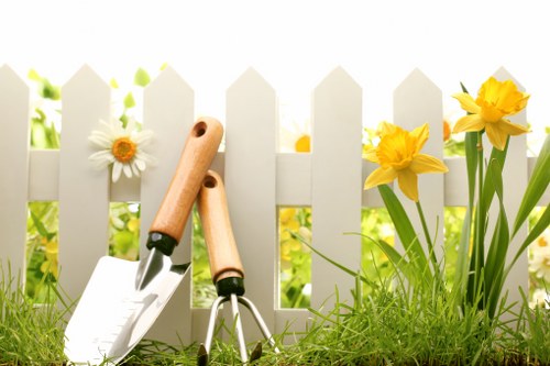 A garden scene featuring a white picket fence in the background with yellow daffodils and white daisies growing along the base. In the foreground, there is well-maintained green grass with a small gardening trowel and a hand fork with wooden handles resting on the grass, indicating active gardening or lawn maintenance. The natural lighting suggests a bright, possibly spring day, highlighting the fresh, vibrant greenery and flowering plants. The setting appears to be a typical residential garden, possibly within the St Johns Wood area, showcasing elements commonly involved in garden renovation and landscaping services offered by Landscapers St Johns Wood. The overall scene emphasizes outdoor gardening tools, diverse plant life, and clean landscaping that aligns with professional garden renovation work in the local area.