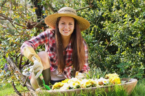 A young woman with long brown hair, wearing a wide-brimmed straw hat, plaid shirt, and gardening gloves, is kneeling in a lush garden during daytime. She is smiling while tending to a compost bin or garden bed filled with vibrant yellow and purple flowering plants. The garden features a variety of green foliage, including shrubs, small trees, and a hedge in the background. The area is well-maintained with neatly arranged flower beds and a healthy, dense lawn with short, even grass. Sunlight filters through the leaves, creating a bright and cheerful outdoor environment, ideal for gardening and lawn care activities. The setting illustrates an outdoor space that could be serviced by professional landscapers in St Johns Wood, focusing on planting, pruning, and general garden maintenance.