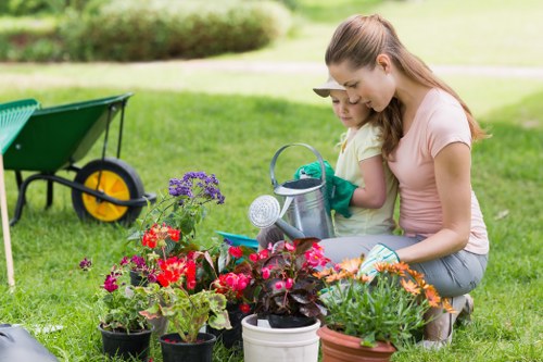 A woman and a young girl are kneeling on a well-maintained grassy garden lawn in a residential outdoor space, engaged in gardening activities. The foreground features a variety of potted plants with vibrant red, purple, and orange flowers, extending across the lush green grass. Behind them, there are garden tools and a watering can placed beside these plants. In the background, the garden includes a green wheelbarrow with black handles and yellow wheels, situated on the grass near a backdrop of neatly trimmed hedges and a lightly blurred lawn area. The scene is captured on a clear day, with natural light illuminating the outdoor space, reflecting typical spring or summer weather conditions. The overall setting suggests a cared-for garden area suitable for minor landscaping and planting work, consistent with gardening services in the St Johns Wood area, with a focus on nurturing flower beds and maintaining lawn health, as provided by Landscapers St Johns Wood.