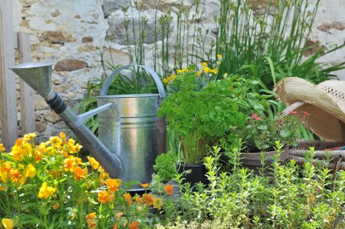 A small, creatively structured garden scene featuring a variety of plants and garden elements. In the foreground, a bed of vibrant yellow and orange flowering plants contrasts with the lush green foliage of nearby herbs and shrubs. Behind these, a weathered metal watering can with a curved handle and spout is positioned next to a collection of potted plants, some with delicate pink flowers. The garden is bordered by a rustic stone wall, adding a natural backdrop to the scene. To the right, a woven straw hat rests on the edge of a wooden garden tool or container, while tall grasses and leafy greenery fill the background, creating a dense, thriving outdoor space. The overall setting appears to be a well-maintained, private backyard or front garden in St Johns Wood, with natural sunlight illuminating the plants and garden tools, emphasizing the textures and vibrant colours that highlight the care taken in garden maintenance and landscaping by local experts such as Landscapers St Johns Wood. This composition offers a detailed view of a lush, productive outdoor garden environment suitable for professional gardening and outdoor upkeep services.