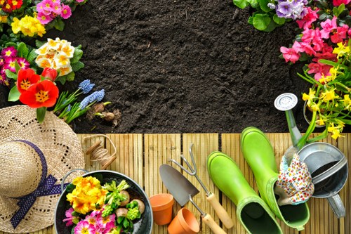 An overhead view of a small, neatly maintained garden area featuring a wooden decking border with various gardening tools and accessories arranged along the edge, including bright green rubber wellington boots, a small watering can, a hand trowel and fork, and a pair of gardening gloves, with flower pots filled with colorful flowering plants such as primroses and pansies positioned at the top left corner. The area is bordered by a lush, healthy hedge and some trees are visible in the background, providing a natural fence and shade, while fresh dark soil remains uncovered, suggesting recent planting or preparation work. The scene is set under natural daylight, with the overall environment appearing tidy and inviting for outdoor gardening activities, indicative of professional or hobbyist gardening efforts in a suburban residential setting typical of St John's Wood.