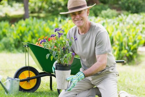 The image depicts a gardener in a light grey t-shirt, beige trousers, and a straw hat, kneeling on a well-maintained lawn in a spacious outdoor garden area during daylight, with soft natural lighting. He is smiling and holding a small potted plant with purple and red flowers, wearing gardening gloves. Behind him, there is a bright green wheelbarrow with large yellow and black wheels, positioned on a lush, dense grass lawn that appears evenly mowed. In the background, there are various trees and shrubs, providing a natural backdrop, with some hedges and flower beds visible at the edges of the garden. The overall scene emphasizes a well-kept outdoor space suitable for gardening and landscaping work, reflecting professional garden maintenance and planting activities that could be offered by local landscapers such as Landscapers St Johns Wood, operating in the NW8 postcode area. The environment looks clean, orderly, and vibrant, highlighting the aesthetic appeal of a landscaped garden setting.
