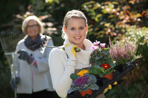 A young woman with blonde hair, smiling and wearing gardening gloves, holds a black tray filled with various potted flowering plants, including pink, red, and purple blossoms, in a well-maintained garden during daytime. The garden features a lush, green lawn in the foreground, bordered by vibrant borders and a variety of shrubs and trees with colorful foliage in the background, indicating a healthy, landscaped outdoor space typical of residential gardens in St Johns Wood. An older woman, also smiling and dressed in light outdoor clothing, stands behind her, holding gardening tools, with sunlight filtering through the trees, creating a bright and inviting outdoor atmosphere suitable for gardening and landscape maintenance services.