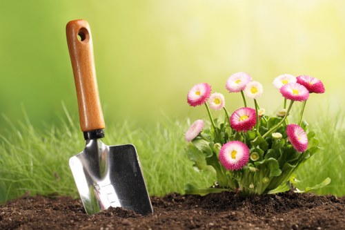 The image depicts a close-up view of a garden bed with freshly turned dark soil, situated on a well-maintained lawn with lush, vibrant green grass. To the right, there is a small cluster of pink and white flowering plants with round blooms, contrasting against the surrounding foliage. In the foreground, a wooden-handled trowel with a metal blade is partially inserted into the soil, indicating ongoing planting or soil preparation work. The background features a softly blurred green gradient, suggesting sunlight filtering through nearby trees or vegetation, creating a bright and welcoming outdoor environment. This scene exemplifies typical gardening activities, such as planting and soil tending, which are essential parts of professional landscaping and lawn care services, including those offered by Landscapers St Johns Wood, serving areas like NW8. The image emphasizes the importance of careful garden layout and healthy plant growth in residential outdoor spaces, aligning naturally with gardening expertise and landscape maintenance in the local London area.