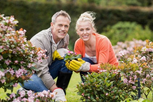 A man and woman are kneeling in a well-maintained garden in St John's Wood, surrounded by flowering shrubs and lush greenery. The man, wearing a light grey shirt, and the woman, dressed in an orange jacket and blue jeans, are smiling and holding a pair of gardening gloves and a small potted plant. The garden features neatly trimmed bushes, vibrant pink and purple flowers, and a backdrop of mature trees and a neatly mowed lawn, indicating regular outdoor maintenance and care. The scene is set on a bright day with natural sunlight illuminating the garden, emphasizing its healthy plants and tidy appearance, reflecting professional gardening and landscaping services provided by Landscapers St Johns Wood. The environment suggests an attractive, landscaped outdoor space suitable for relaxation or family enjoyment, demonstrating the kind of garden work the company offers in the local area near postcode NW8, London.